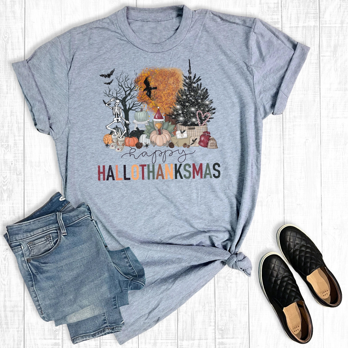 Gray t-shirt with 'Happy HALLOTHANKSMAS' design, paired with jeans and shoes on a wooden surface.