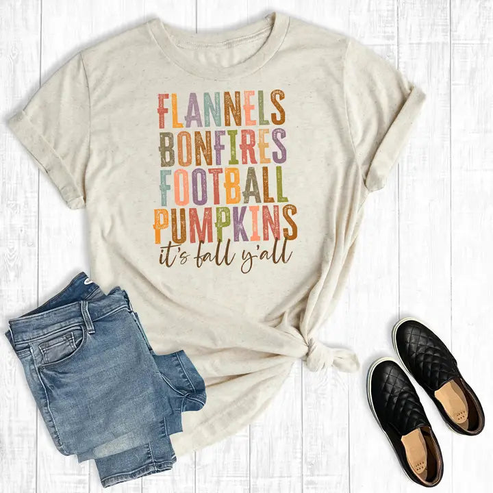 Beige t-shirt with colorful fall-themed text, paired with jeans and shoes on a wooden surface.