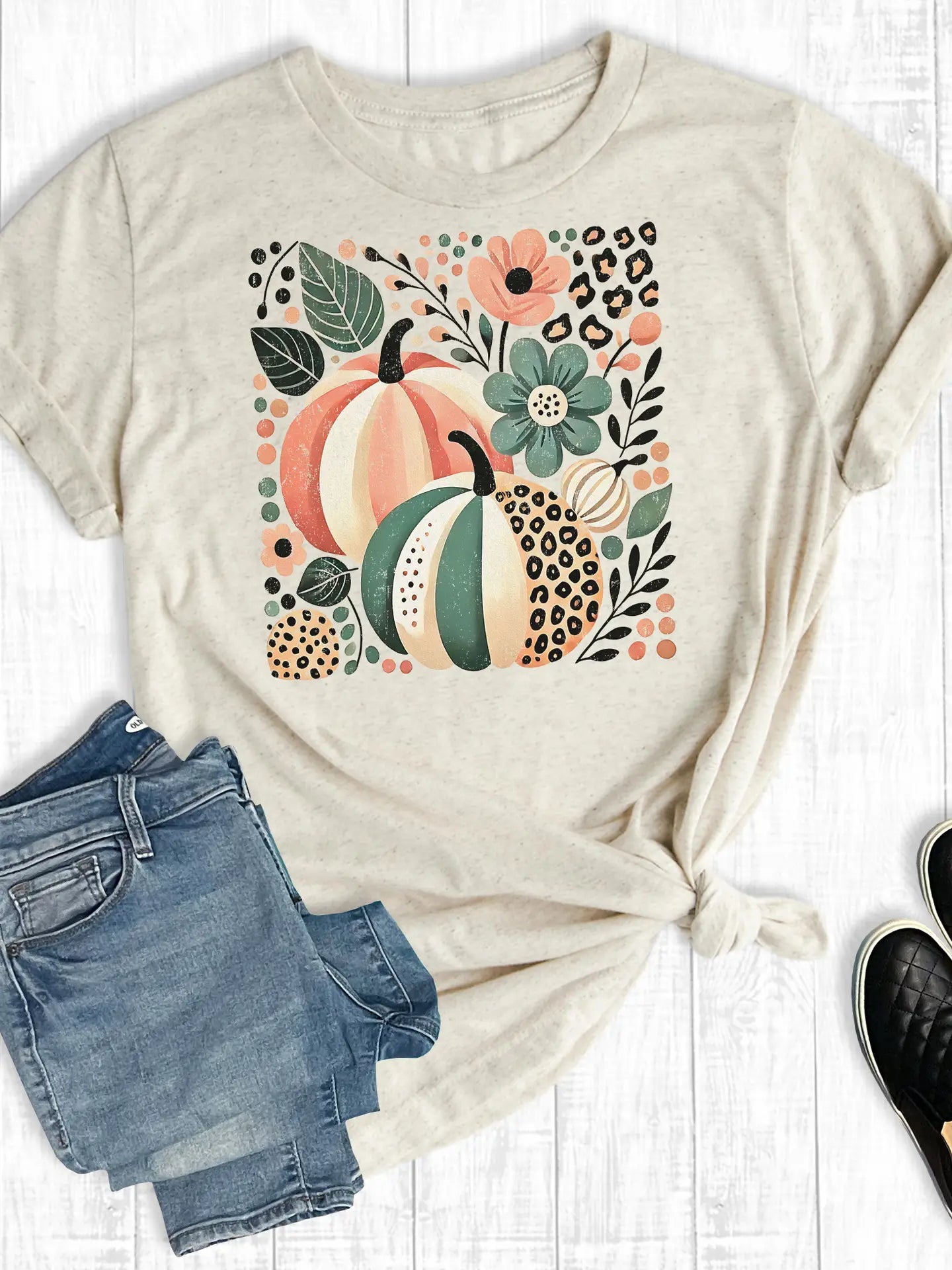 Beige t-shirt with pumpkin and floral design on a white surface with jeans and shoes.