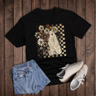 Black t-shirt with a graphic design on a wooden surface, accompanied by denim shorts and white sneakers.