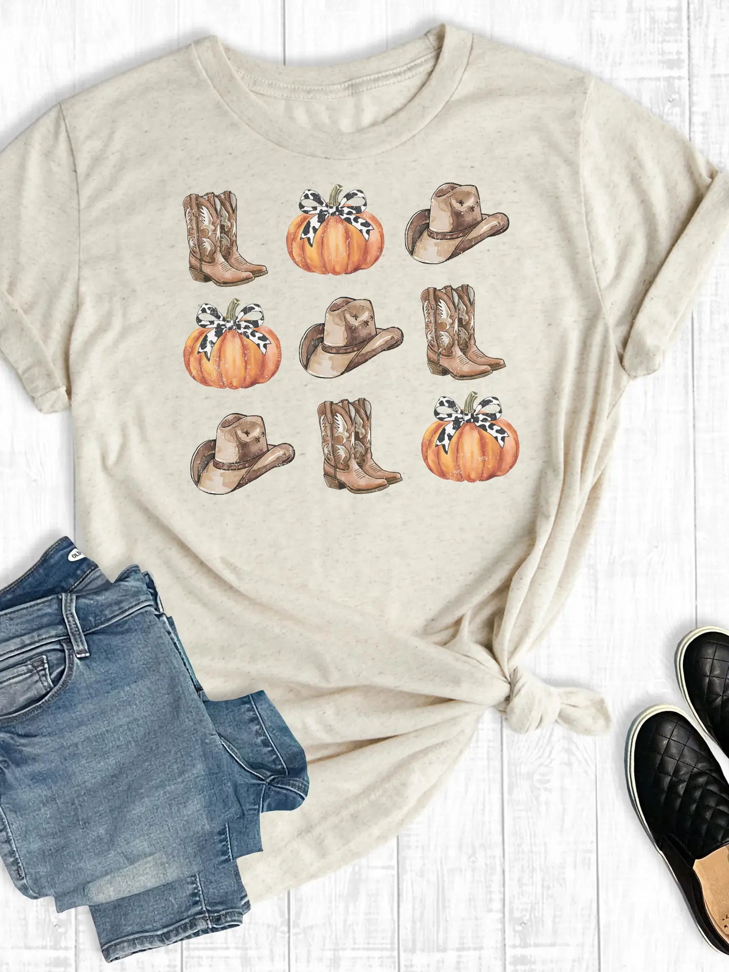 Beige t-shirt with cowboy boots and pumpkins design, paired with denim shorts and black shoes on a white wooden surface.