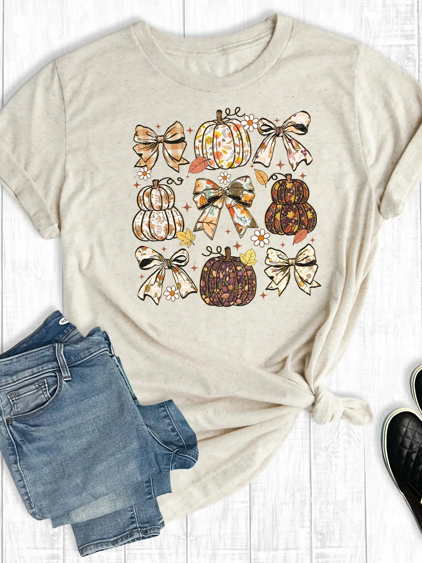 Beige t-shirt with pumpkins and bows design, paired with blue jeans and black shoes on a white wooden surface.